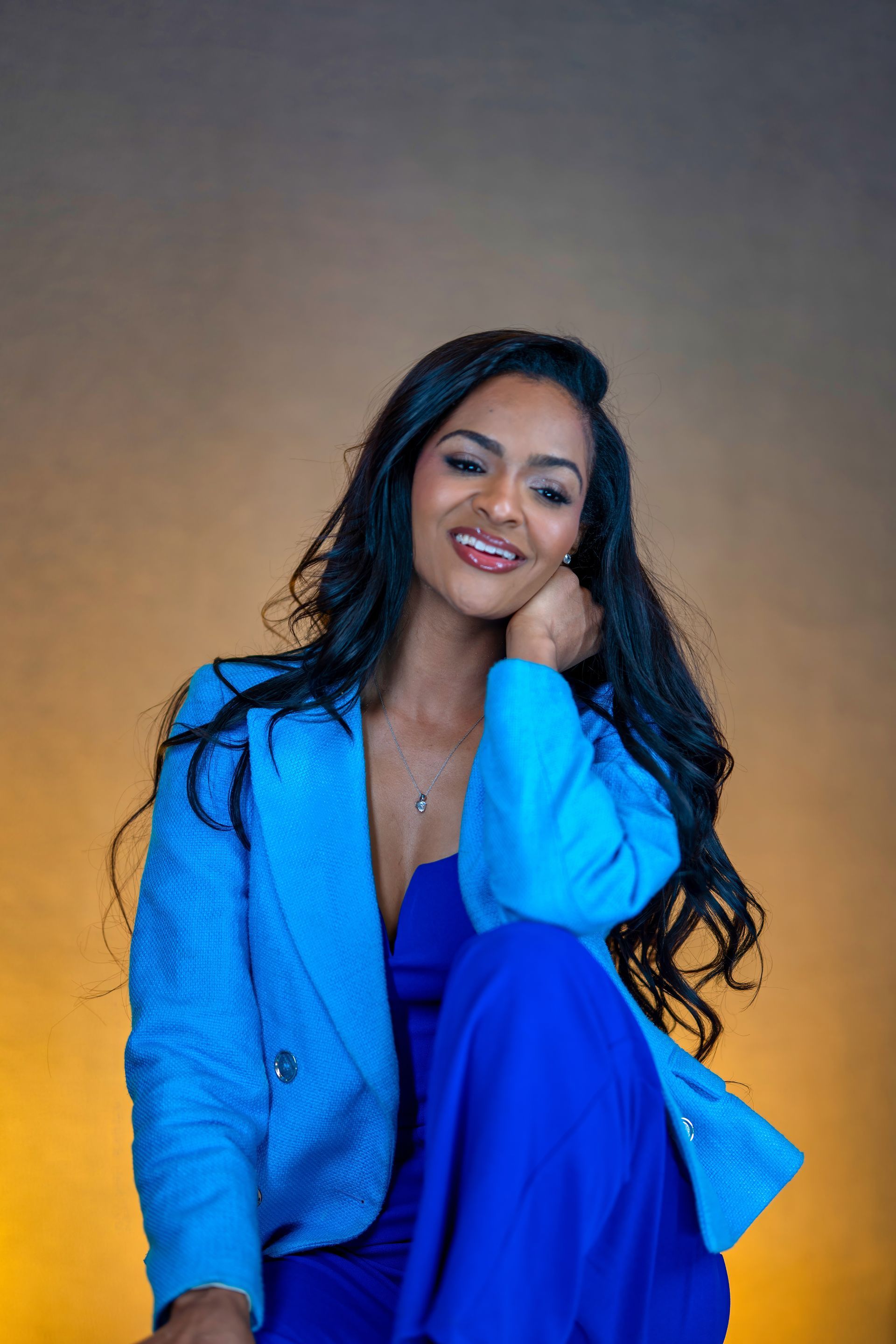 Smiling person in a bright blue blazer and dress, seated against a plain brown background.