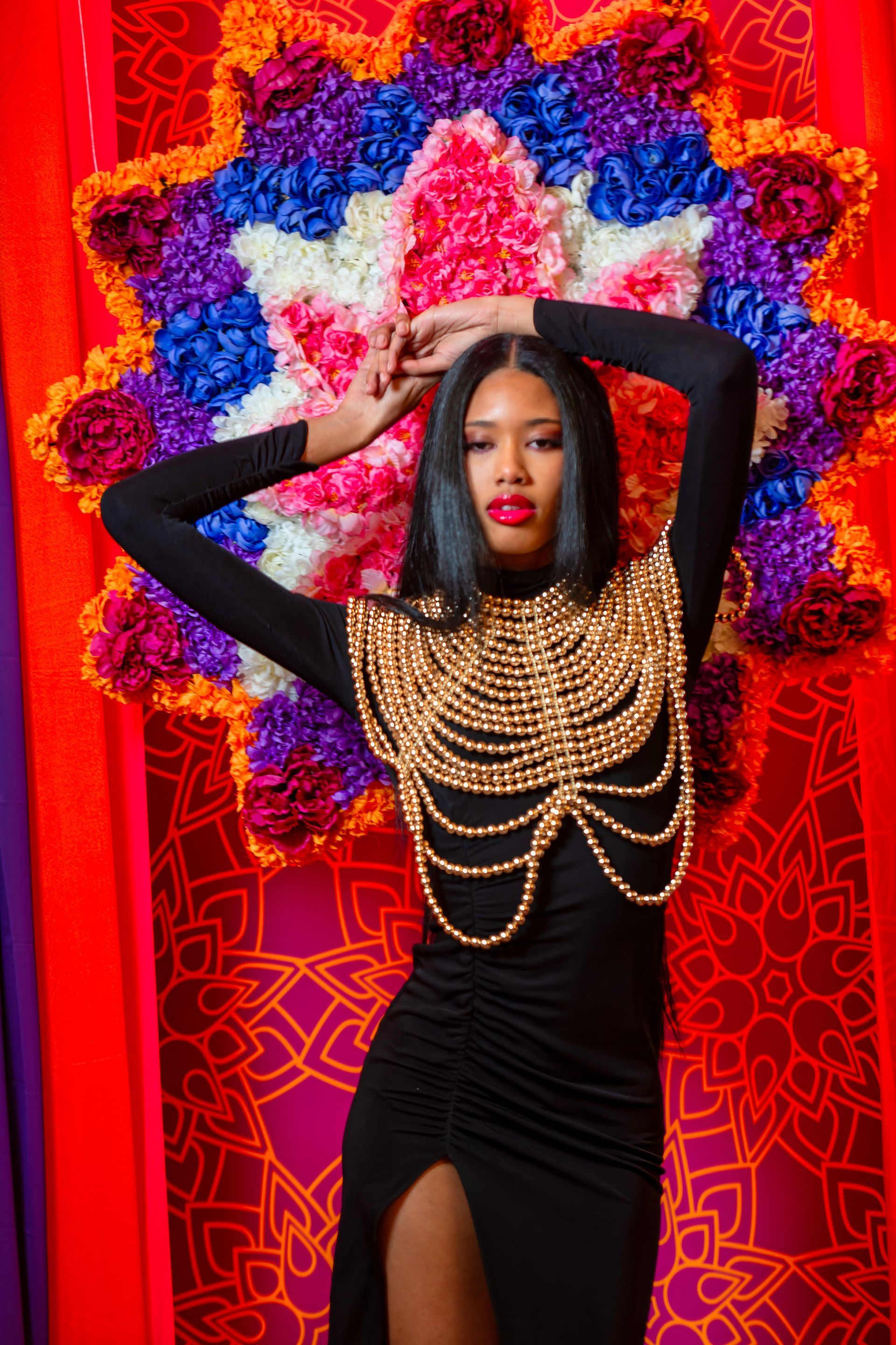 Person in a black dress with gold chains poses against a bright red and floral backdrop.