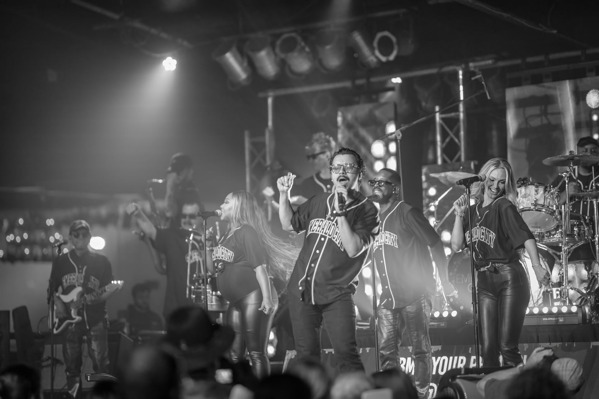Band performing live on stage under bright spotlights, with singer at center and crowd in foreground