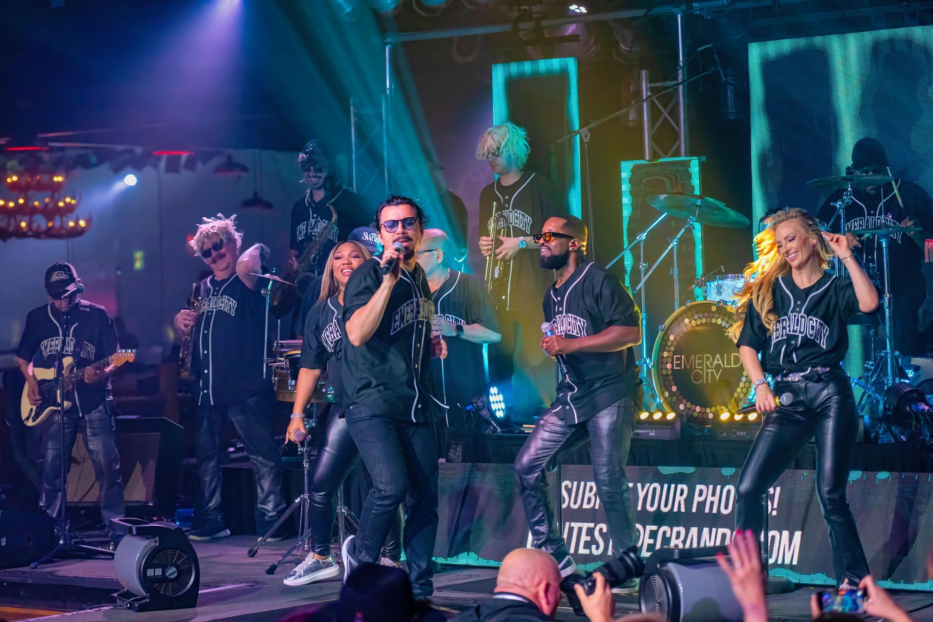 Band performing live on a neon-lit stage, with singer at center and musicians playing guitars and drums.