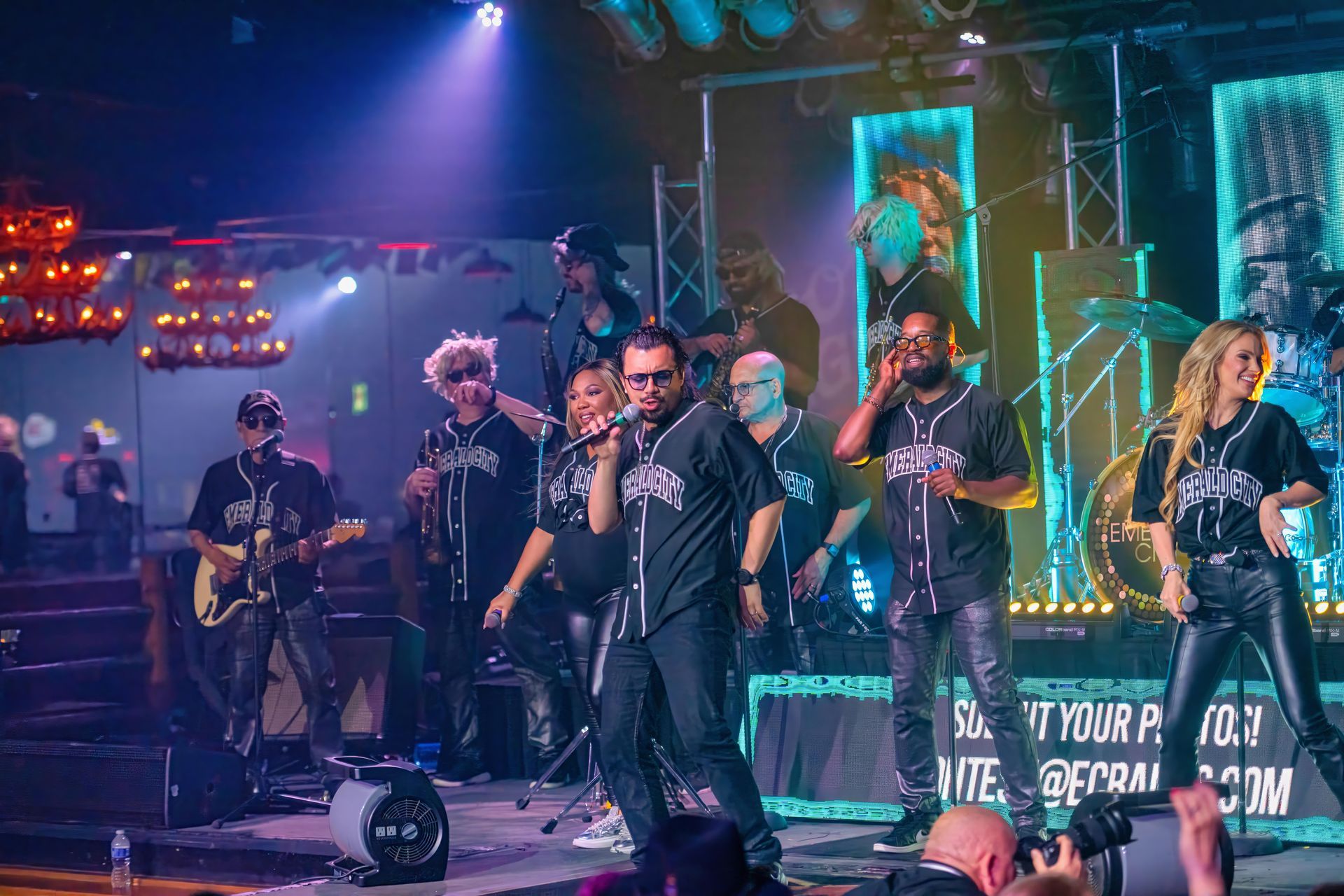 Band performing live on a brightly lit stage with singers, musicians, and colorful neon lighting.