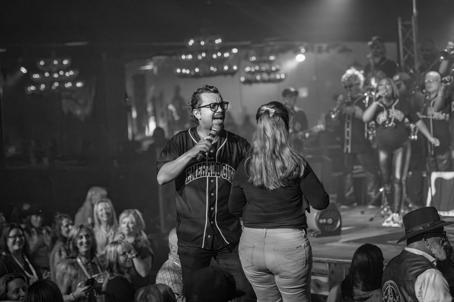 Singer speaking to a woman onstage in a black-and-white concert scene with a cheering crowd