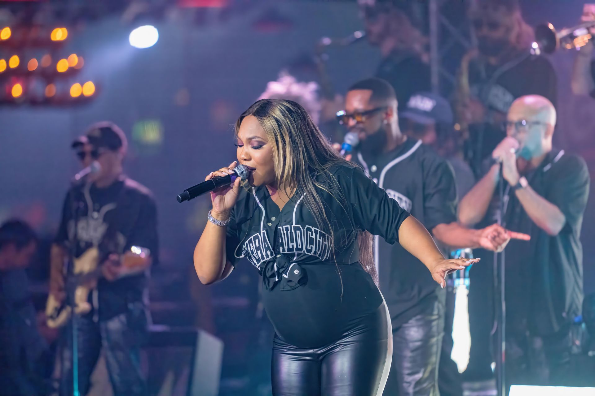Singer performing on stage with backup singers, holding a microphone under colorful concert lights.