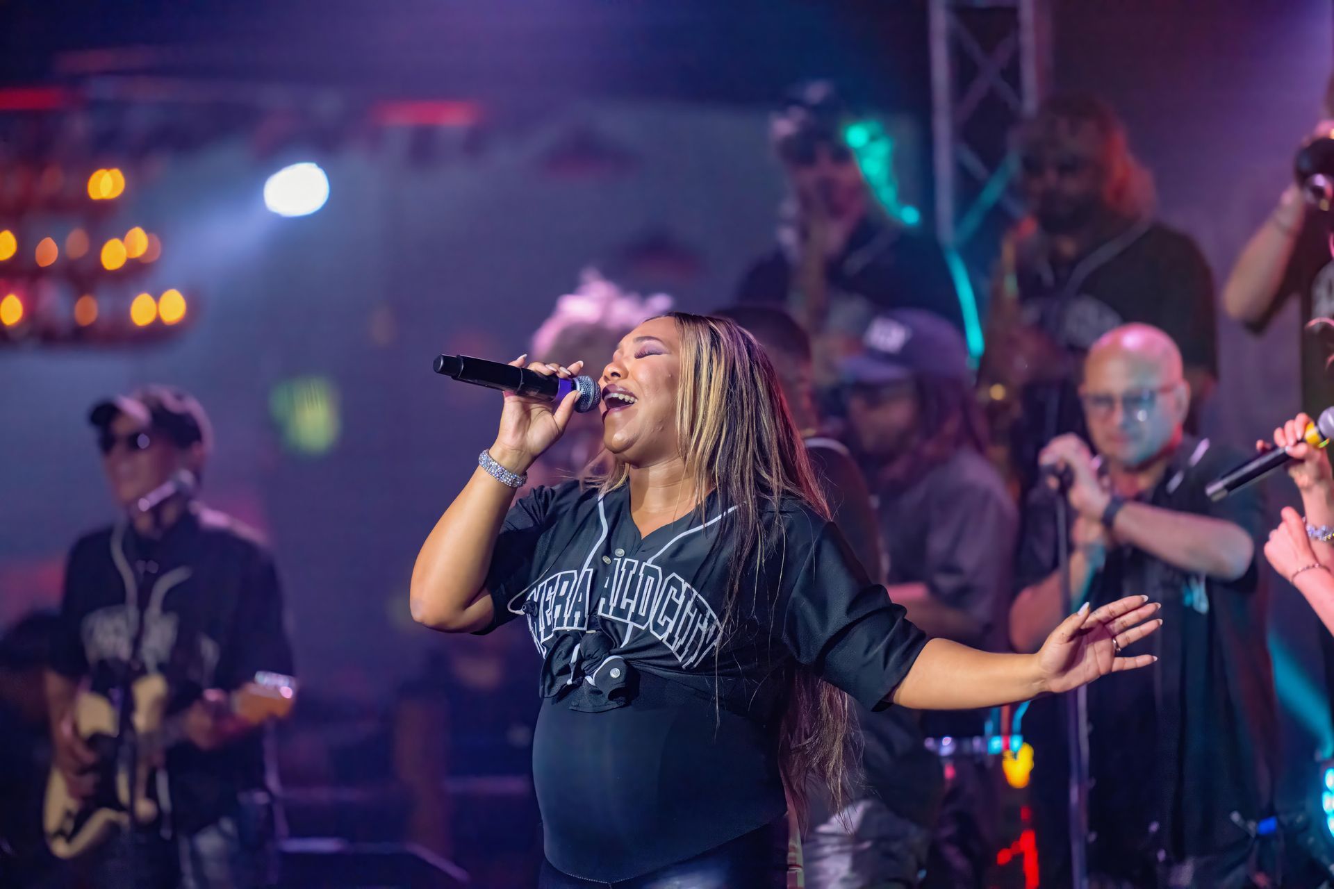 Singer performing on stage with microphone, band visible behind her under colorful concert lights