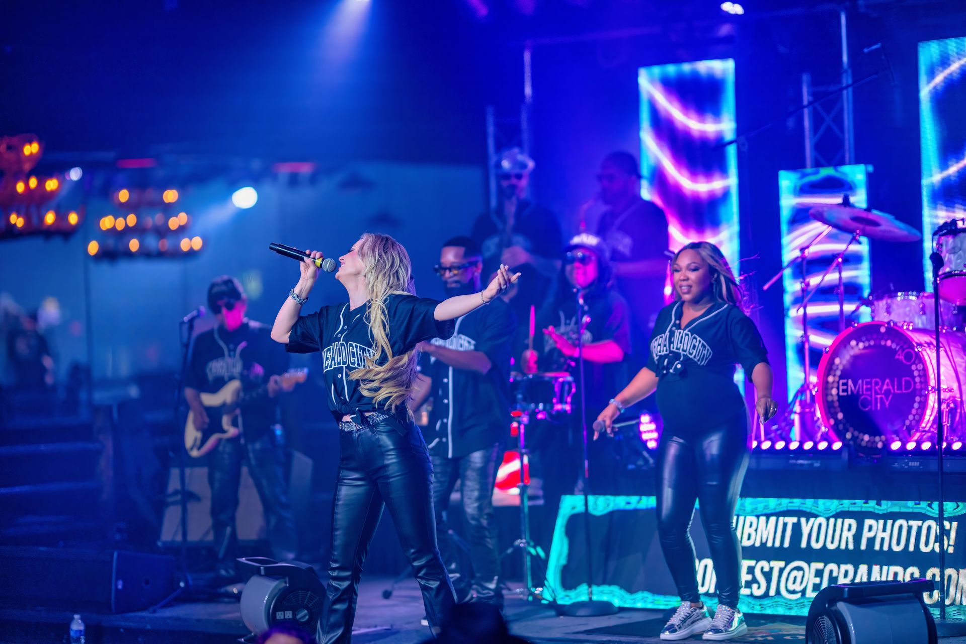 Live band performing on a neon-lit stage, with singers and musicians under blue and purple lights.