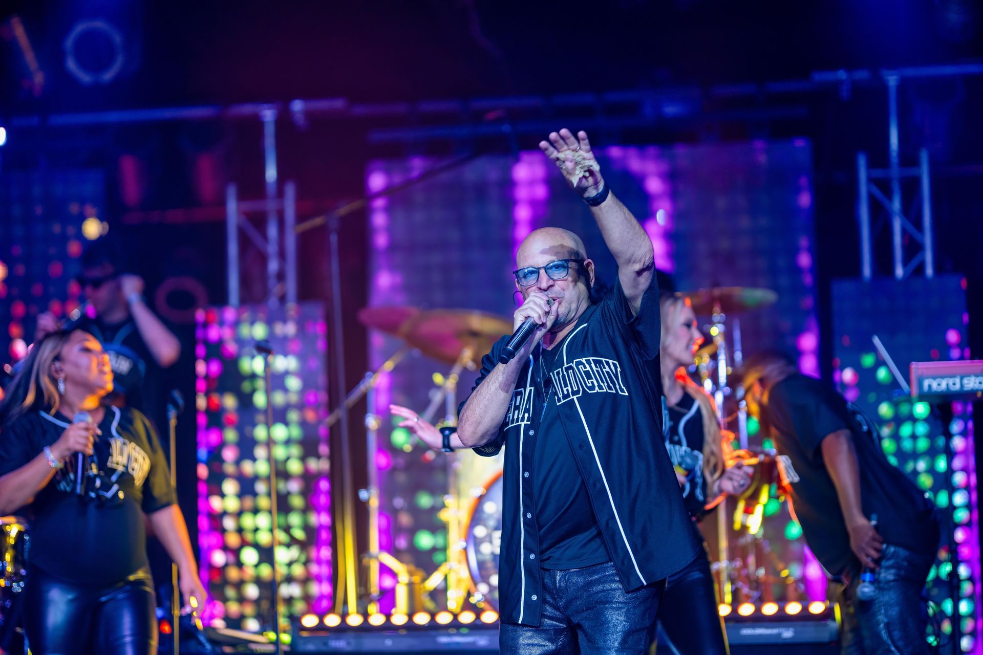 Singer performing on a neon-lit stage, holding a mic and raising one hand, with backup performers behind him.