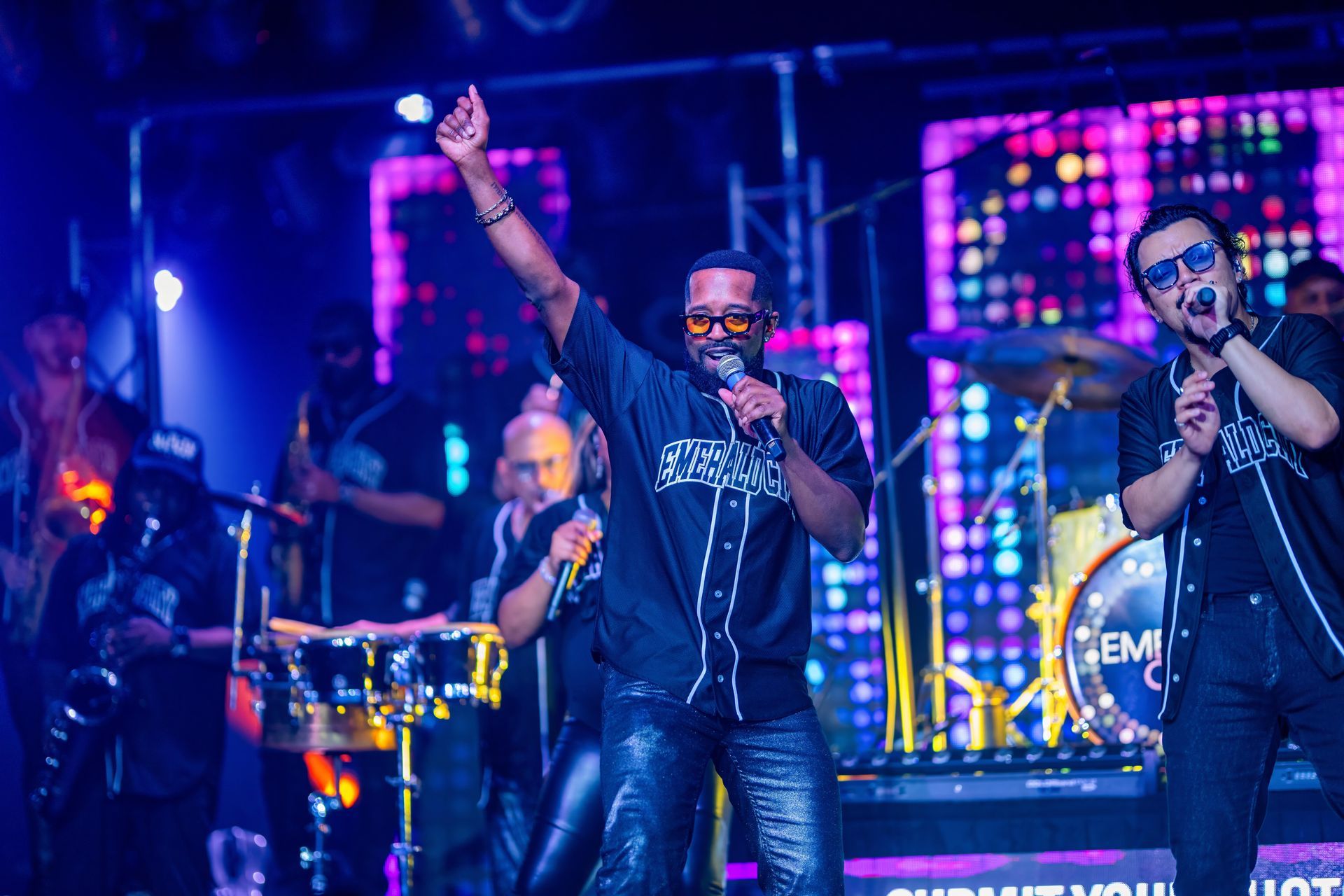 Singer performing on a brightly lit stage with backup musicians and colorful LED lights