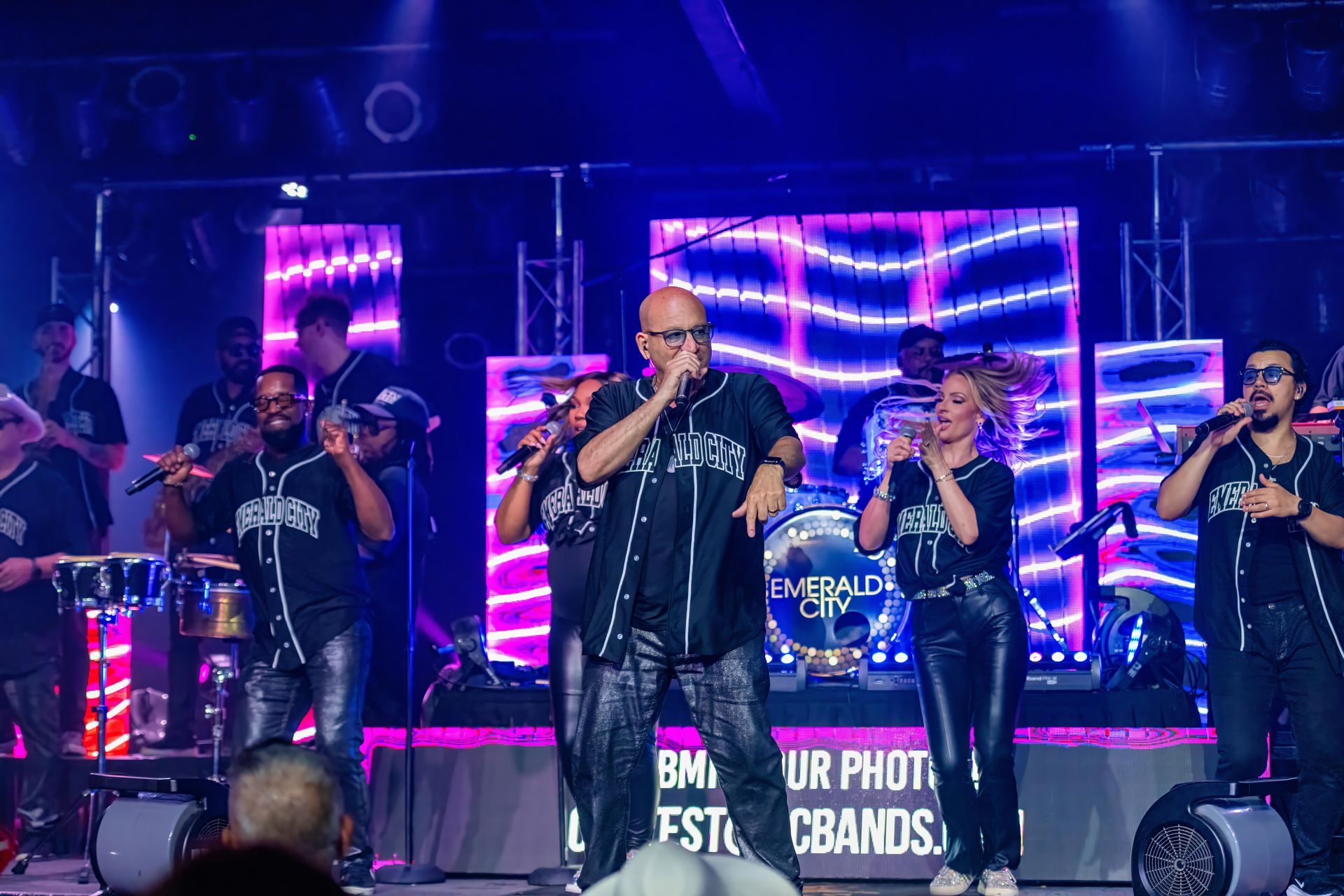 Four singers performing on a neon-lit stage with microphones and LED screens behind them