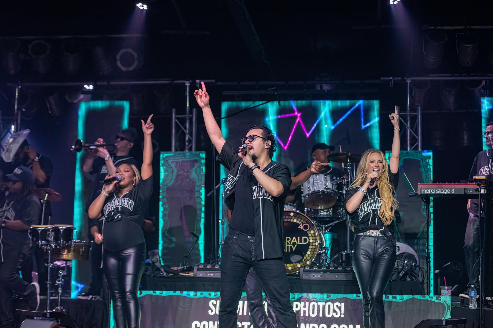 Four performers onstage in black outfits, singing and raising their arms under neon lights