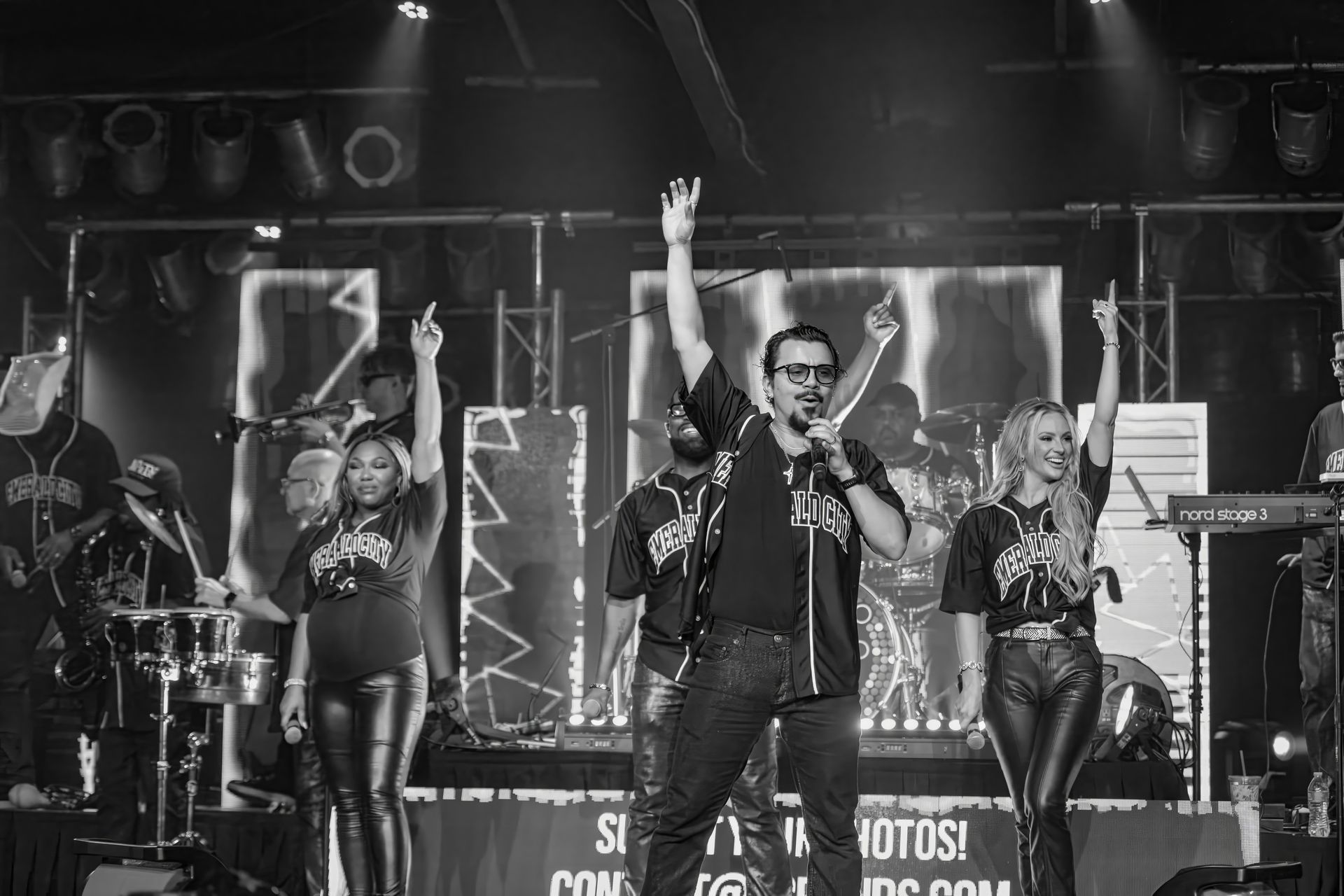 Black-and-white concert scene with three performers on stage, arms raised under bright lights