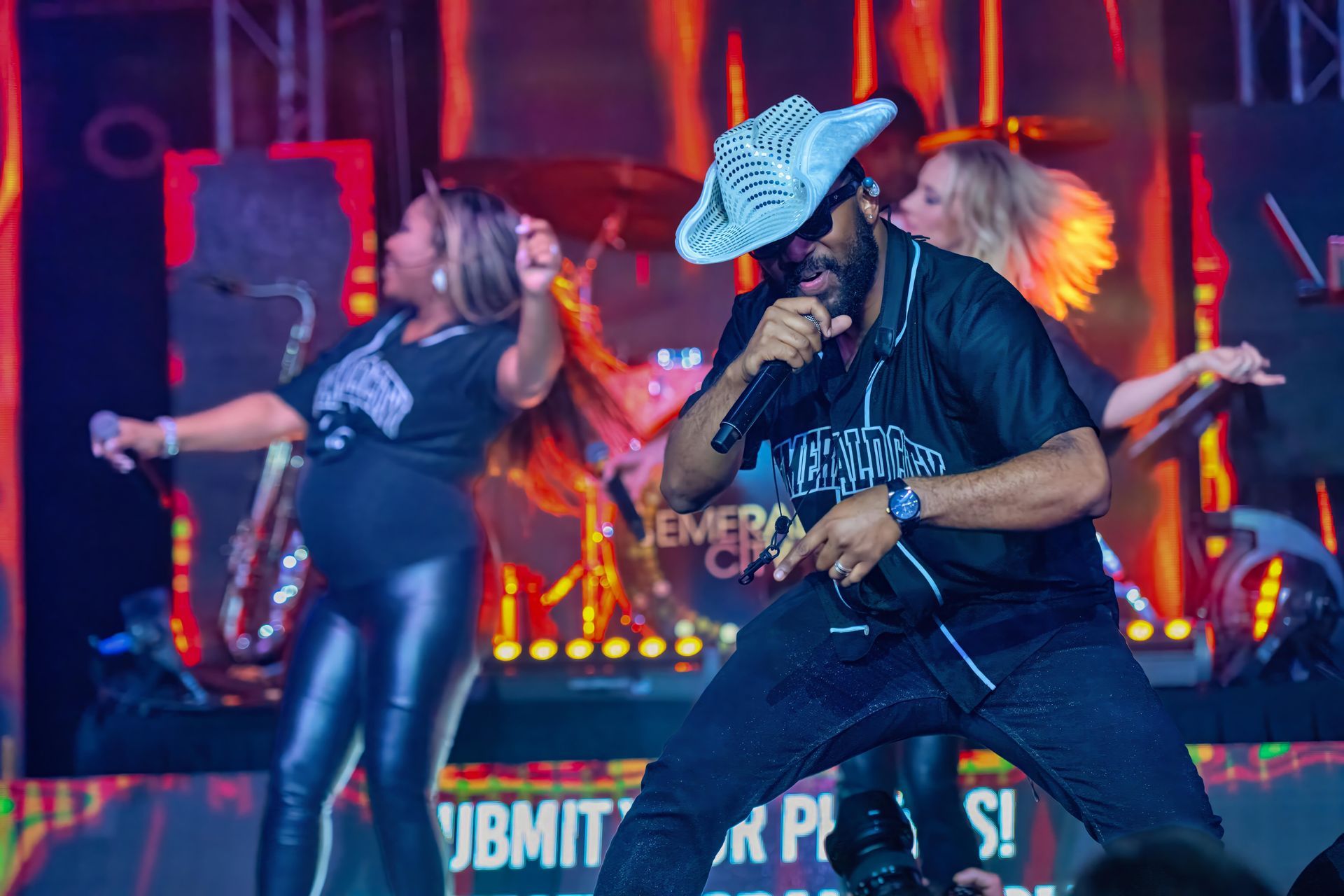 Two musicians performing onstage under red lights, one singing into a microphone while another dances nearby