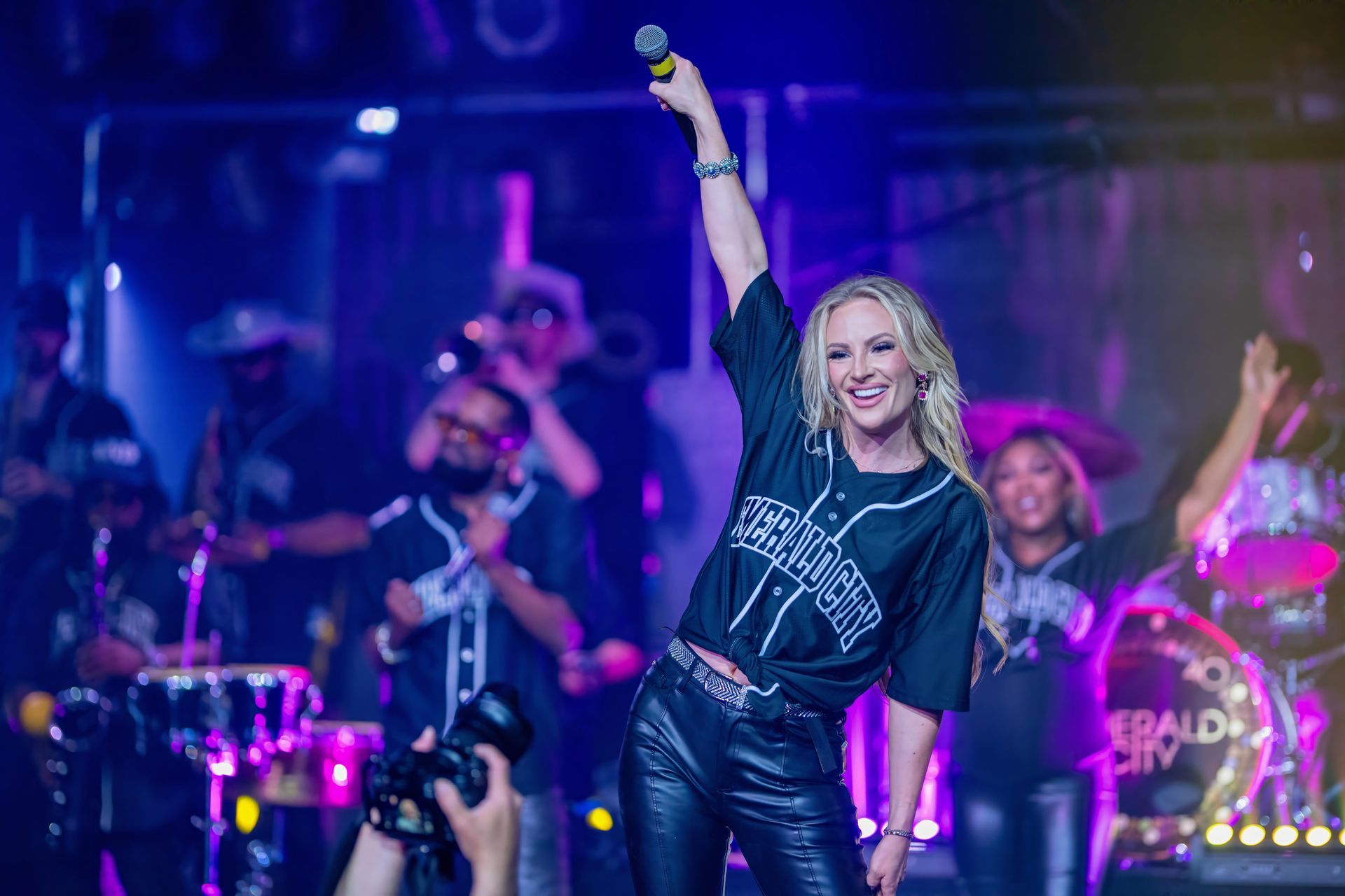 Singer performing onstage with microphone raised, band playing in colorful concert lights