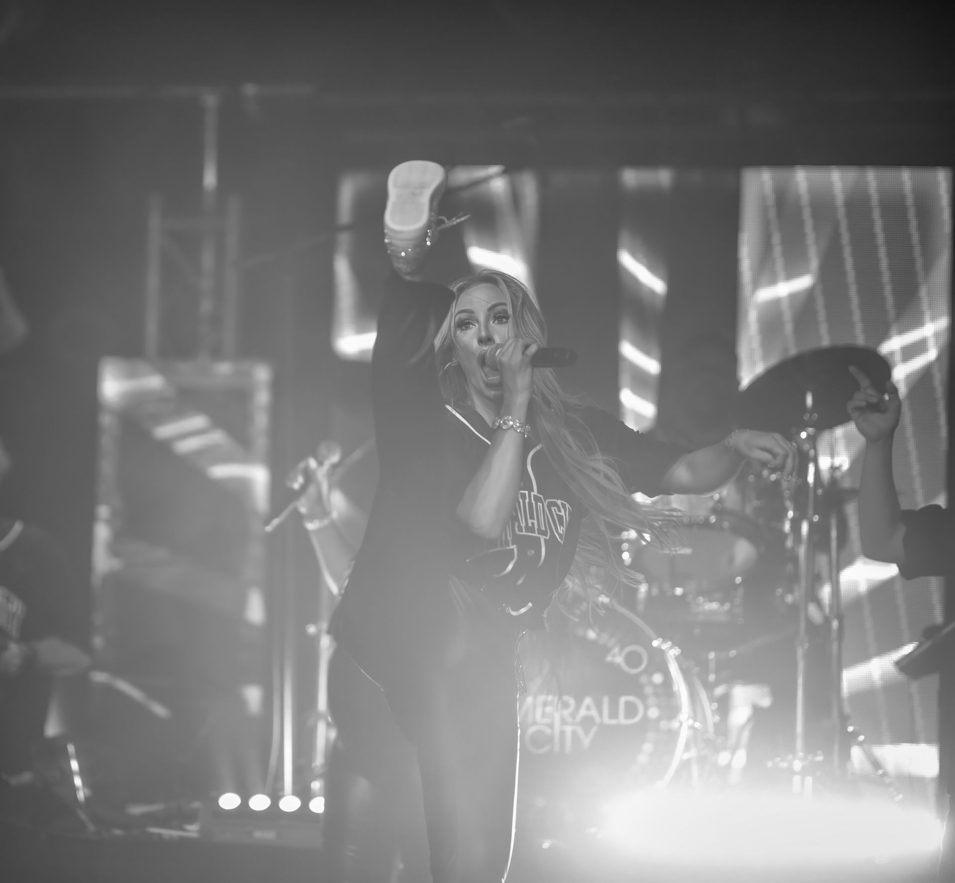 Singer performing on a smoky stage in black and white, with drum kit and bright spotlights behind.