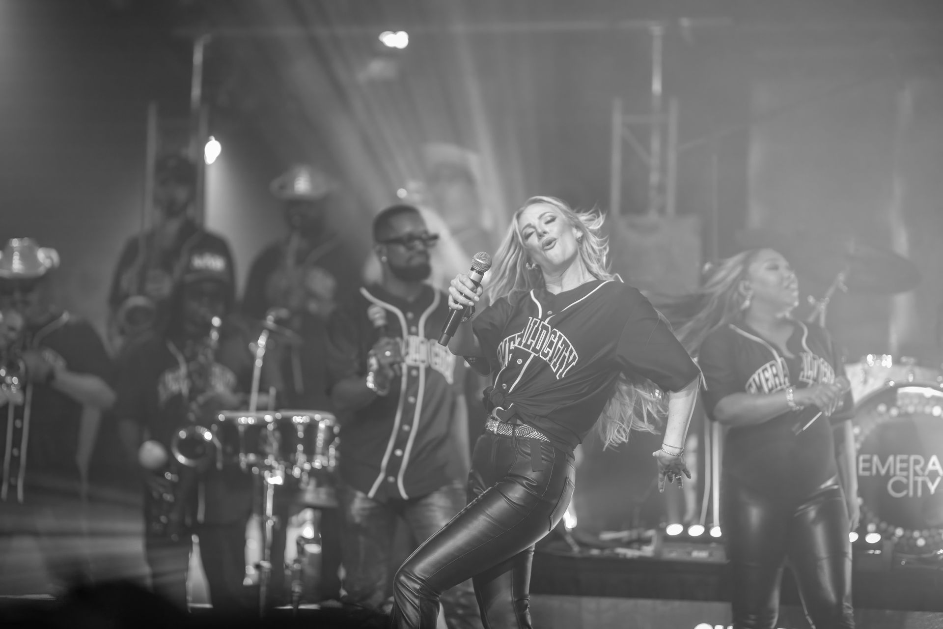 Singer performing on a stage with backup performers and drums under dramatic spotlights