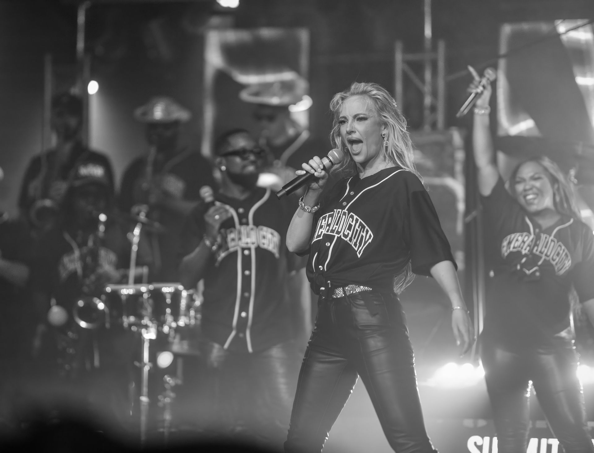 Black-and-white concert scene with a singer holding a microphone center stage and backup performers behind her