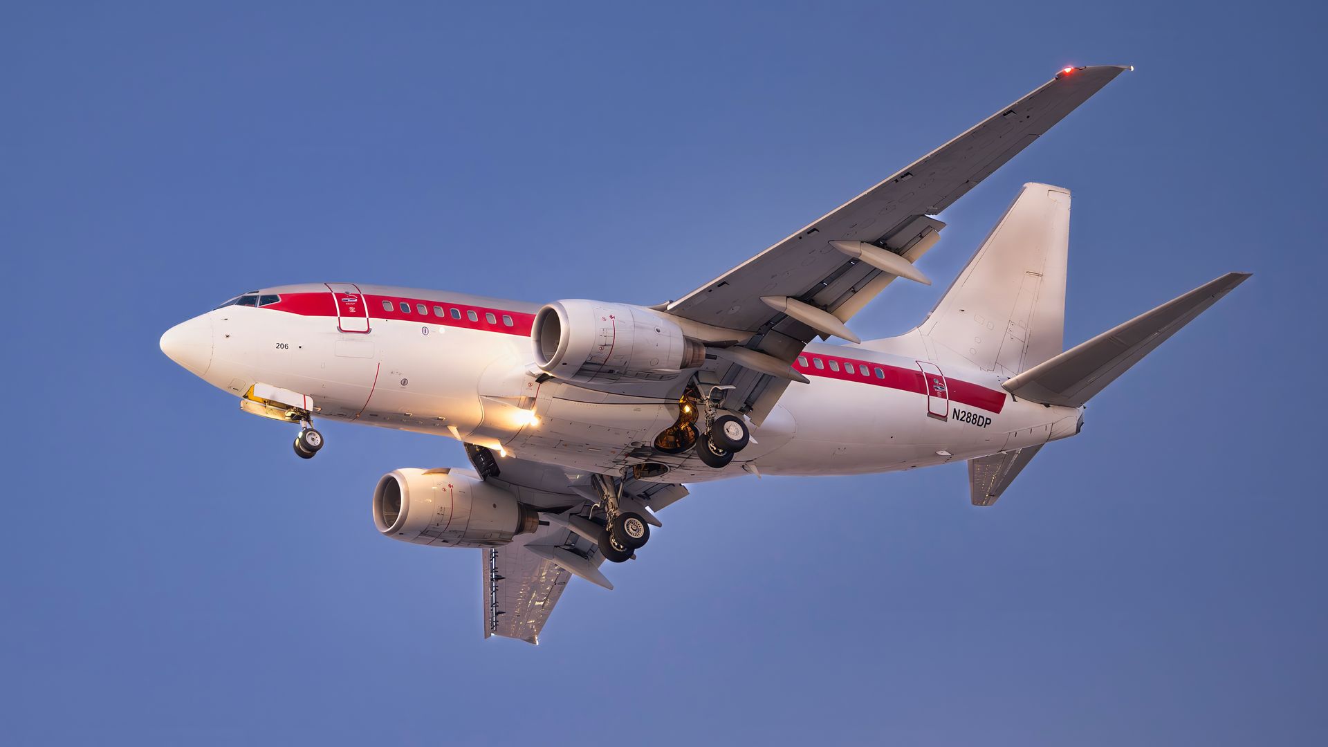A white airplane with a red stripe along its side flies against a clear blue sky, its landing gear deployed.