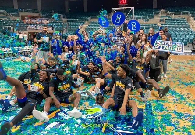 Large team group photo on a confetti-covered basketball court, posing with championship signs and trophy.