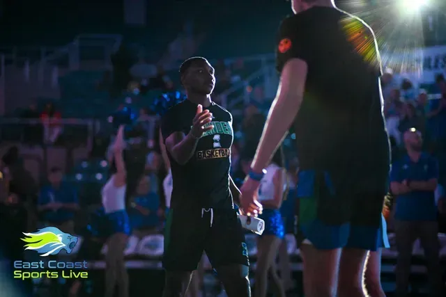 Two athletes in black jerseys on a lit basketball court, with a crowd in the background and one holding a water bottle.