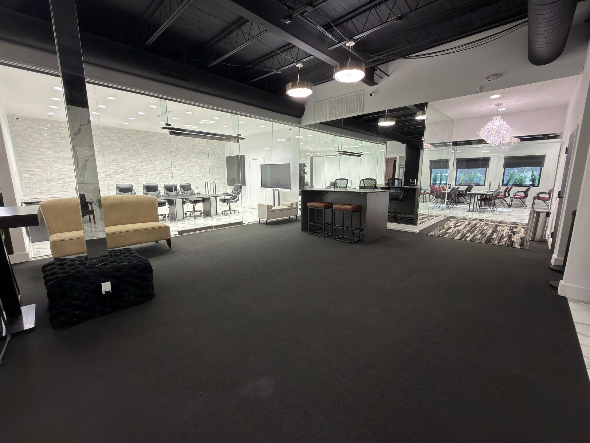 Empty open office/lounge with dark carpet, white walls, tables, chairs, and exposed ceiling lights