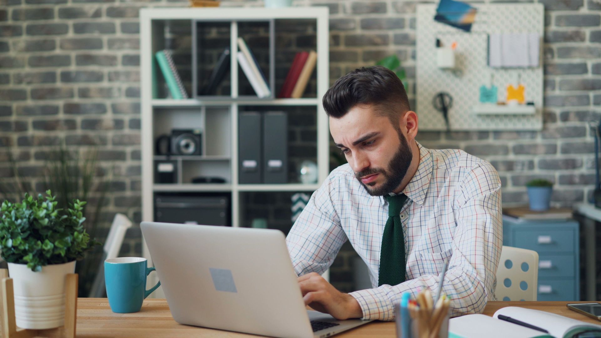 Man working on a laptop at a desk in an office with shelves and plants behind him