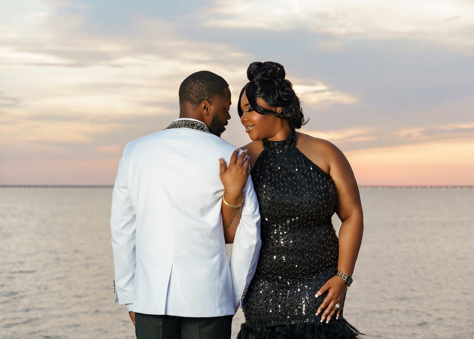 Couple, man in white jacket, woman in black sequin dress, embrace by water at sunset.
