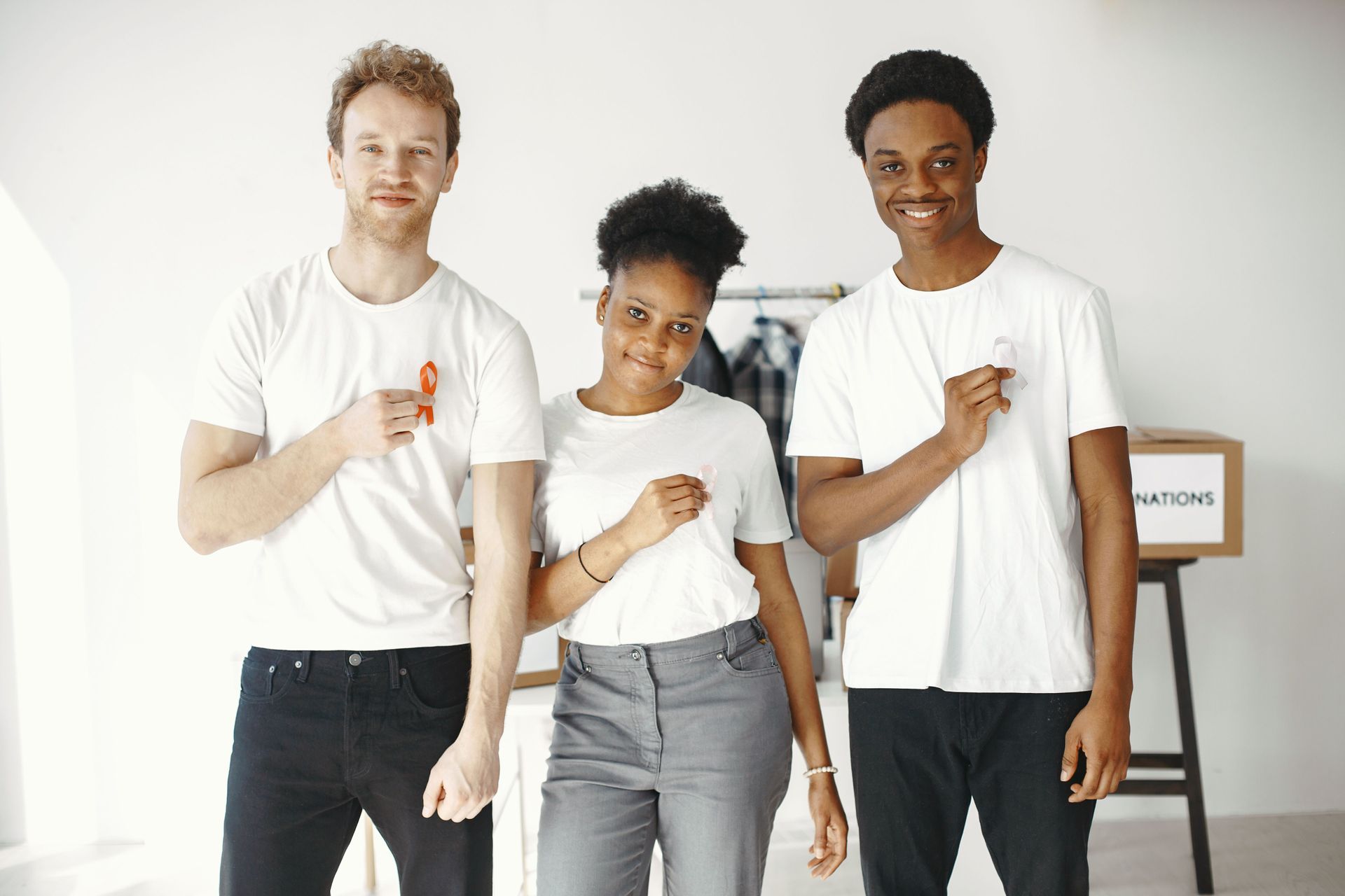 Three people in white shirts stand indoors, each pointing to their chest against a bright wall.