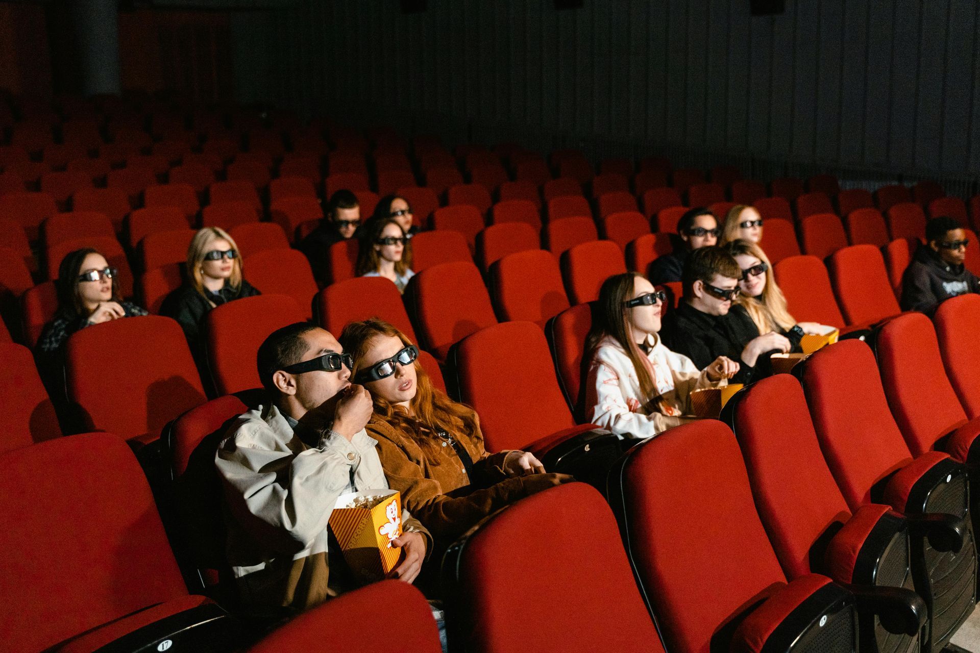 People wearing 3D glasses watch a movie in a red-seated theater. Some eat popcorn.