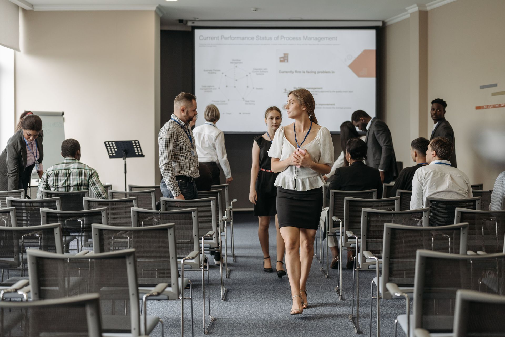 Conference room with people seated and chatting while a woman walks toward the front screen