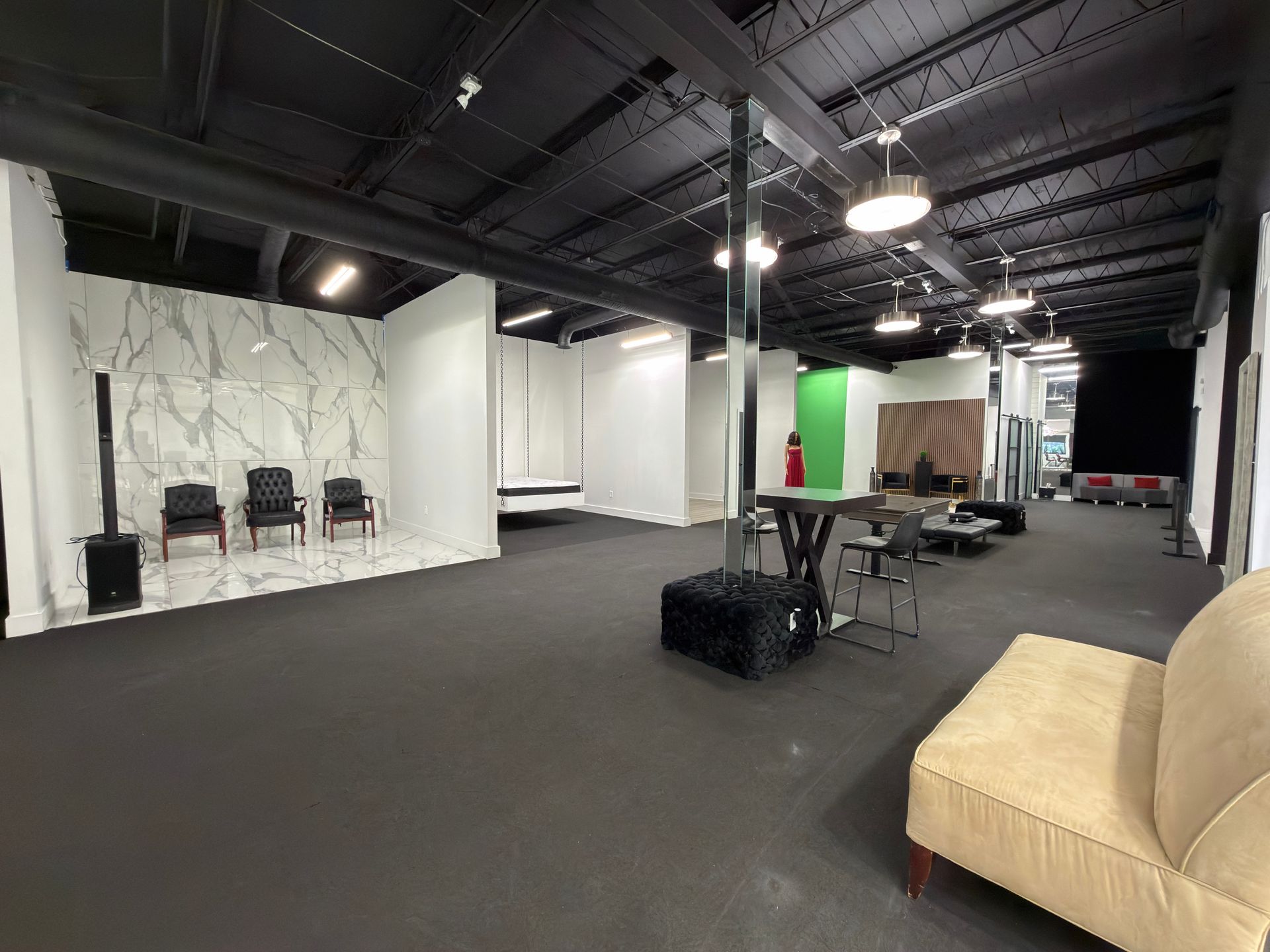 Empty industrial-style studio with black carpet, white walls, chairs, tables, and a beige couch