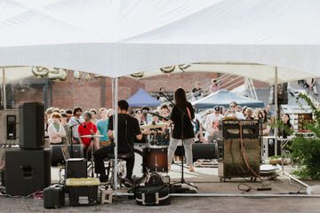 Live outdoor music stage under a white canopy, with performers and equipment in front of a crowd