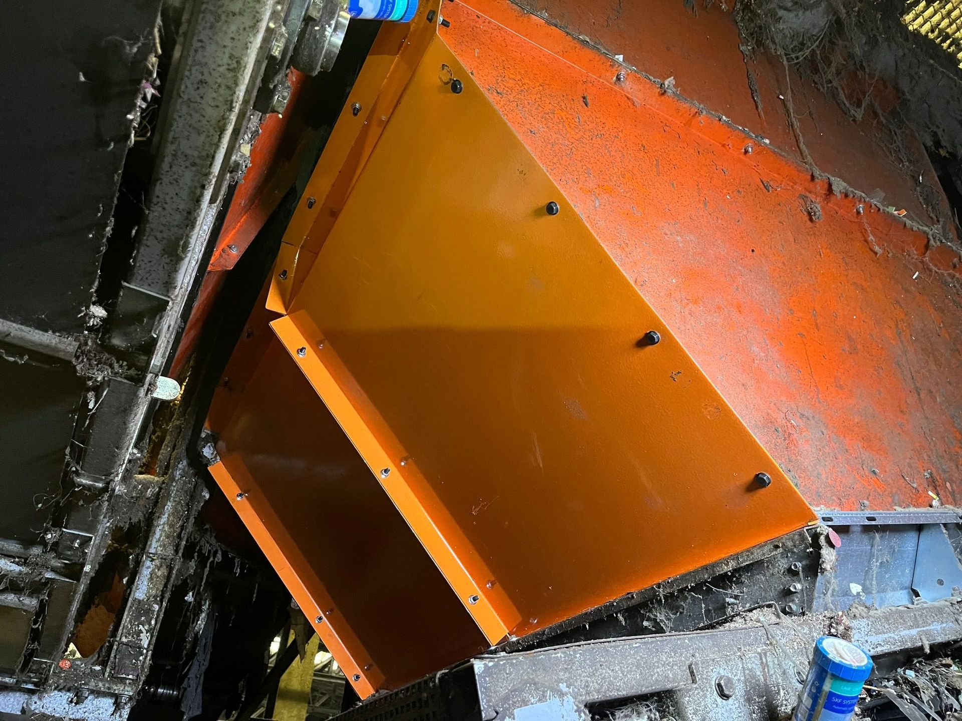 Een oranje metalen stortkoker geplaatst in een machine en bevestigd met schroeven, in een stoffige, industriële omgeving.