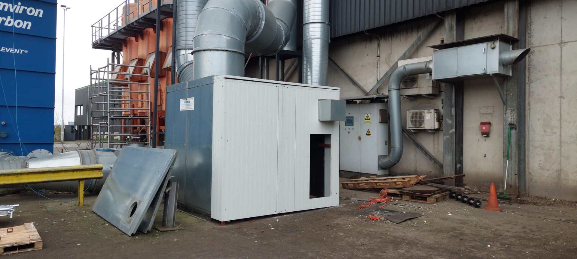 Buitenzijde van een industrieel gebouw met metalen ventilatiesystemen en apparatuur. Er ligt wat bouwafval op de grond.