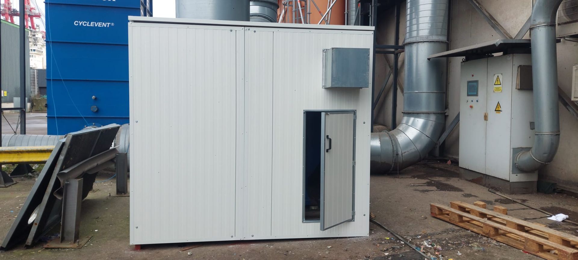 Wit industrieel gebouw met een open deur naast een metalen ventilatierooster en een blauwe container.