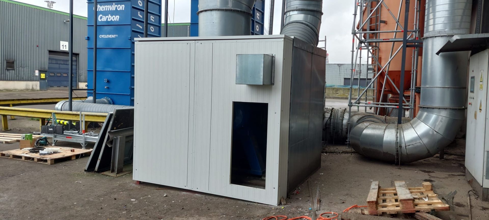 Een wit industrieel gebouw met een rechthoekige opening, metalen buizen en een blauwe container op de achtergrond.