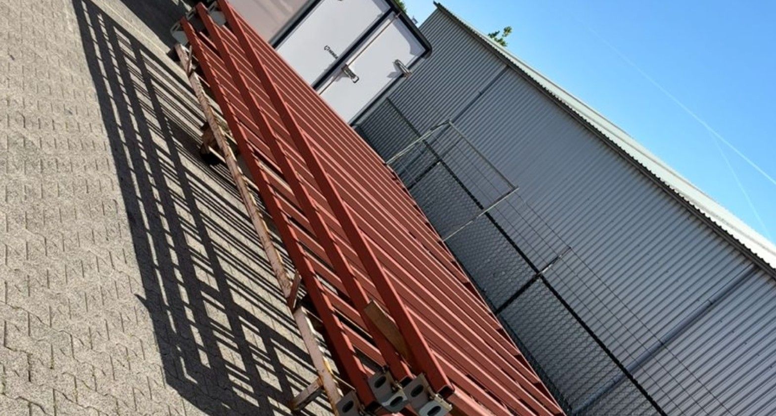 Rode metalen steigers tegen een grijs gebouw, die een lange schaduw werpen op een muur met structuur onder een blauwe hemel.