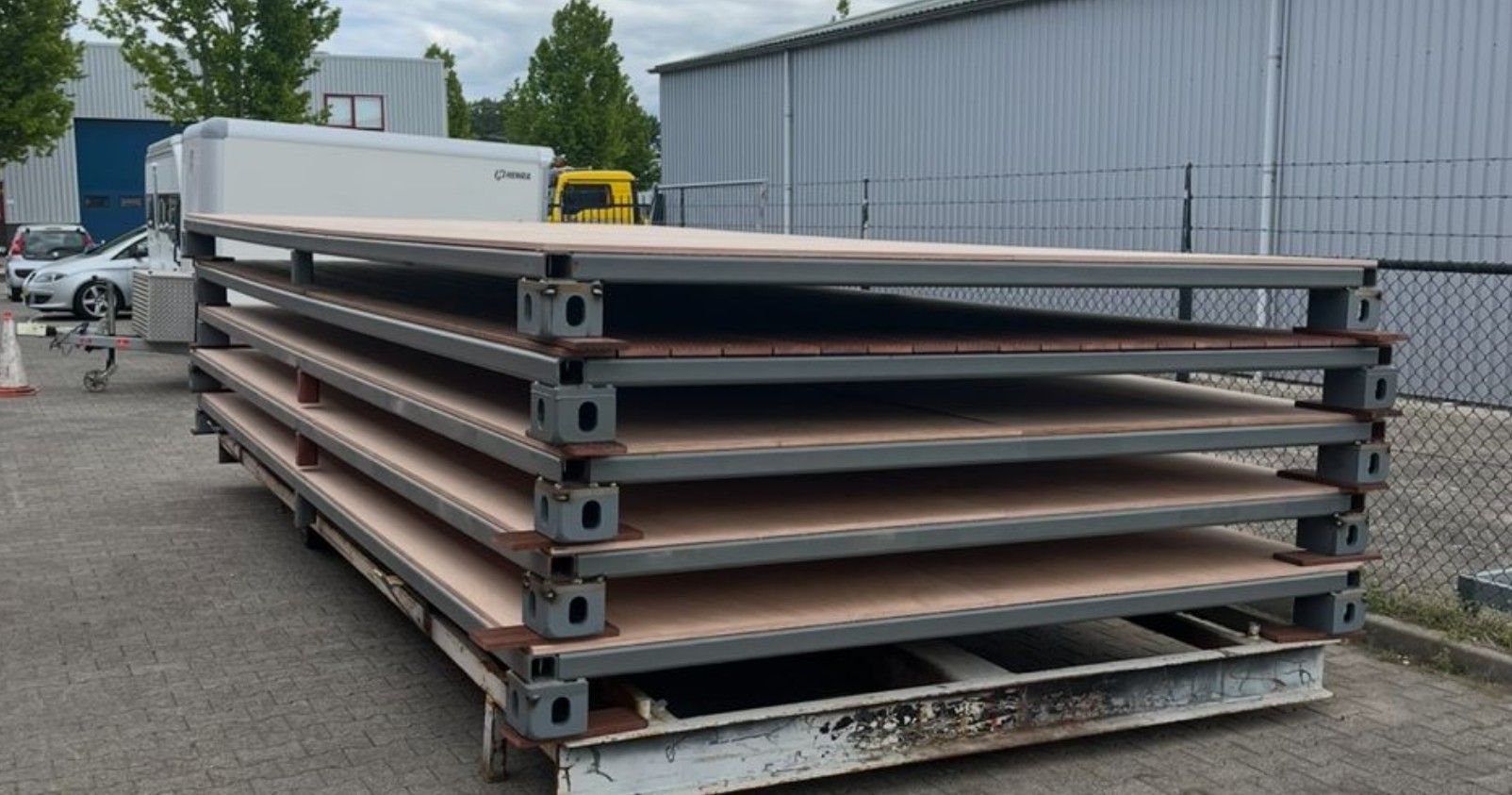 Stapel grote, rechthoekige houten platforms met metalen frames, buiten in de buurt van industriële gebouwen.