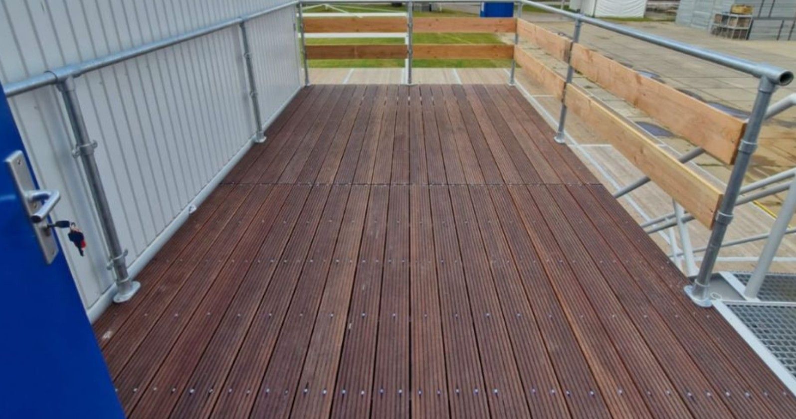 Houten terras met veiligheidsleuningen naast een wit en blauw gebouw.
