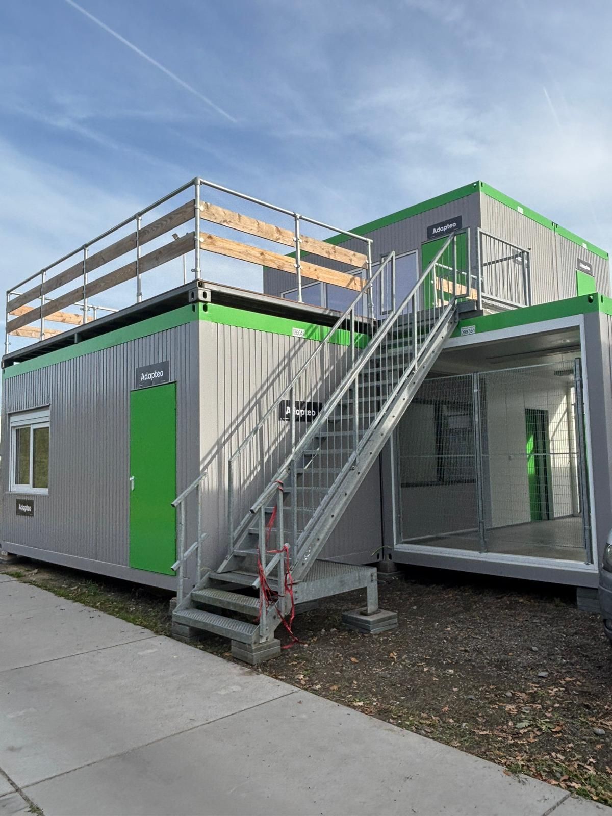 Modulaire constructie met twee verdiepingen en een trap. Zilverkleurige buitenkant, groene accenten en een dakterras.