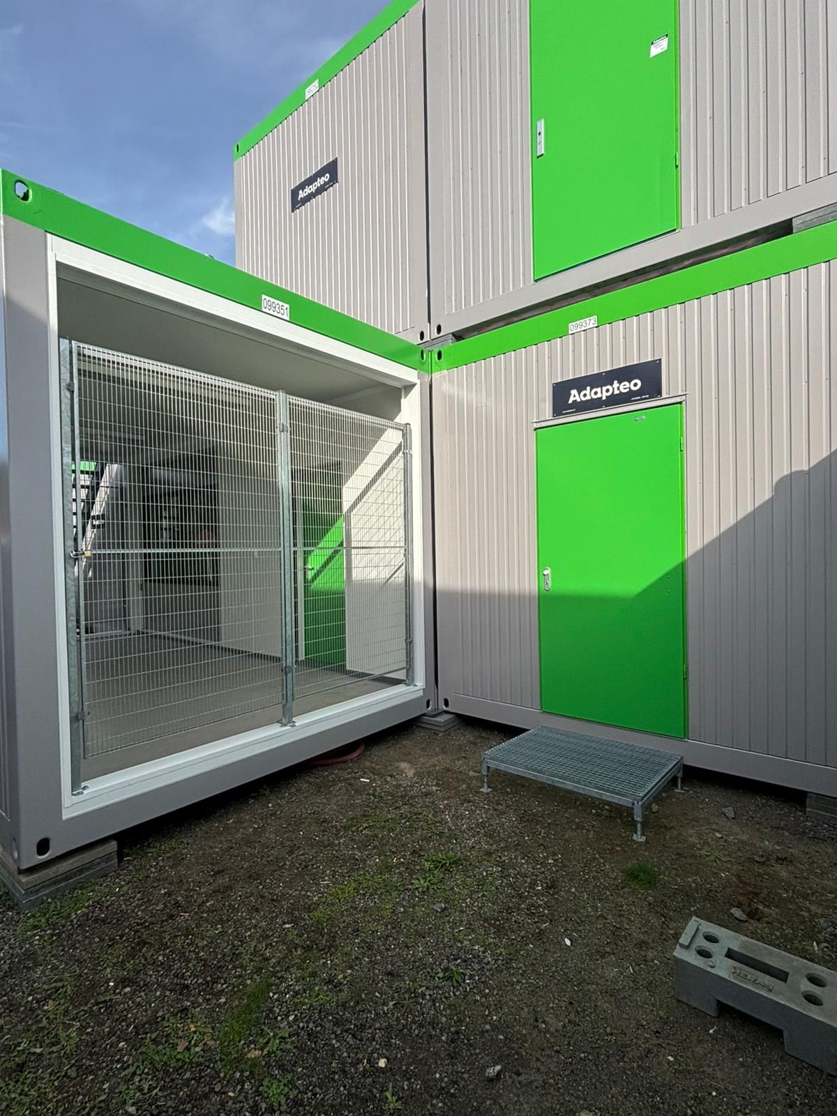 Stapel grijze en groene zeecontainers, waarvan één open is met een metalen rooster en groene deuren.