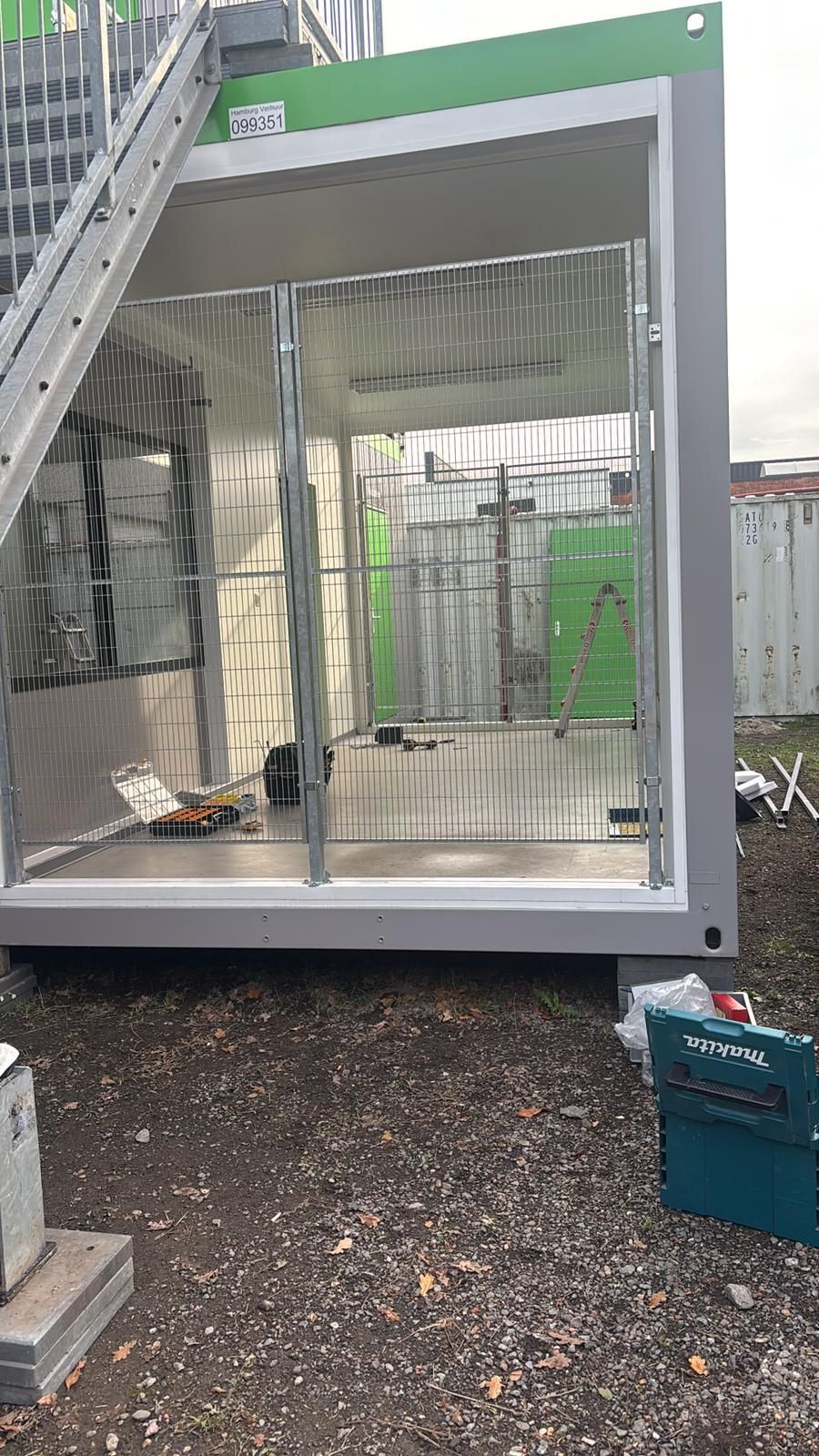 Een containerconstructie met metalen poorten, een ladder en gereedschap aan de buitenkant; het geheel is een bouwplaats.