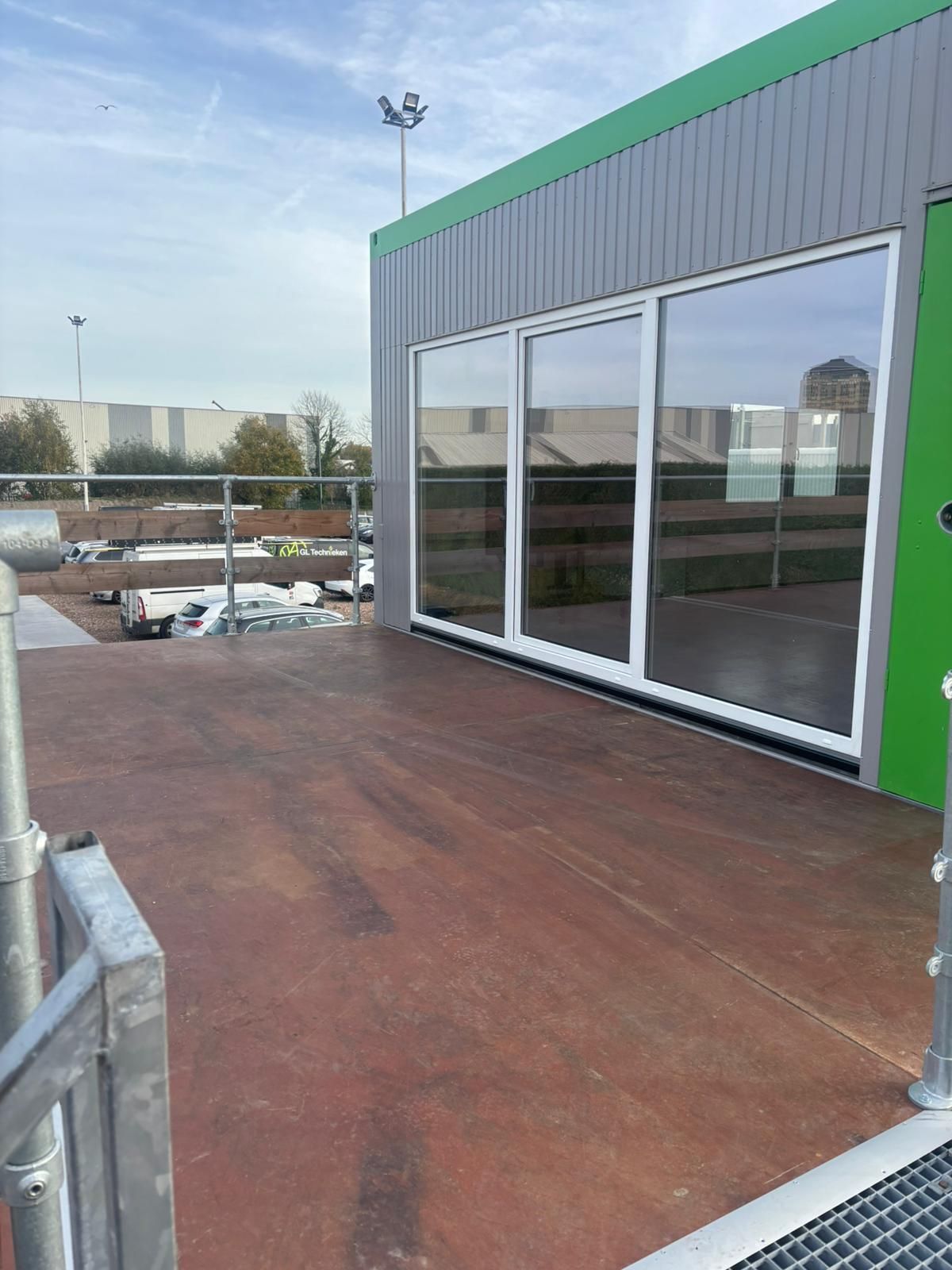 De buitenkant van een gebouw met een groot glazen raam, een bruin houten terras en een metalen balustrade.