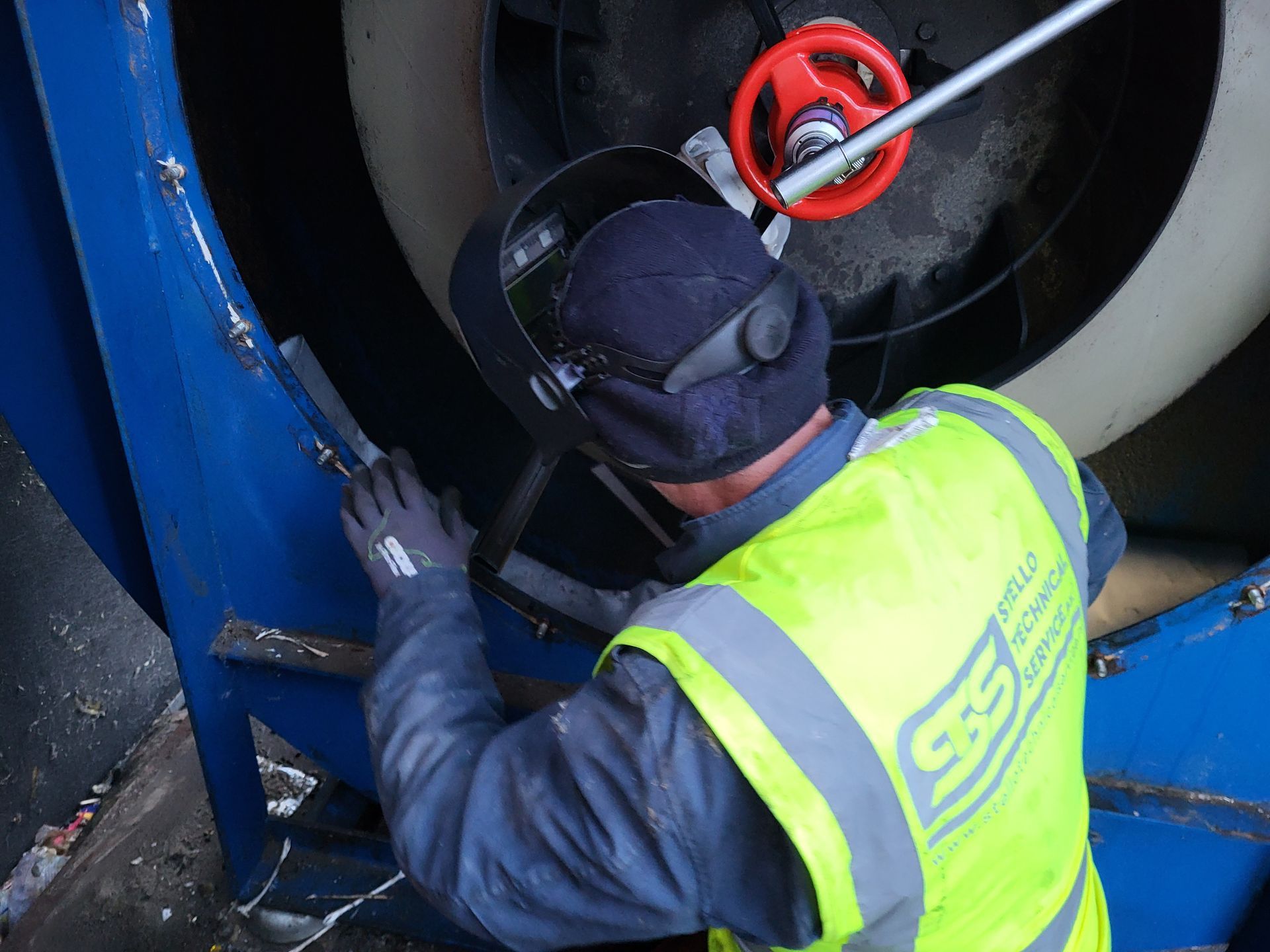 Een persoon die een metalen constructie last, met veiligheidshelm en -vest aan, werkt aan een grote ventilator.
