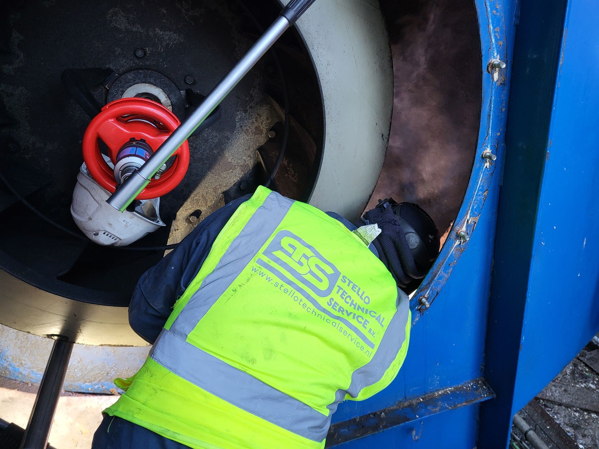 Een persoon in een geel veiligheidsvest inspecteert een machine met een gereedschap. De apparatuur is blauw en zwart.