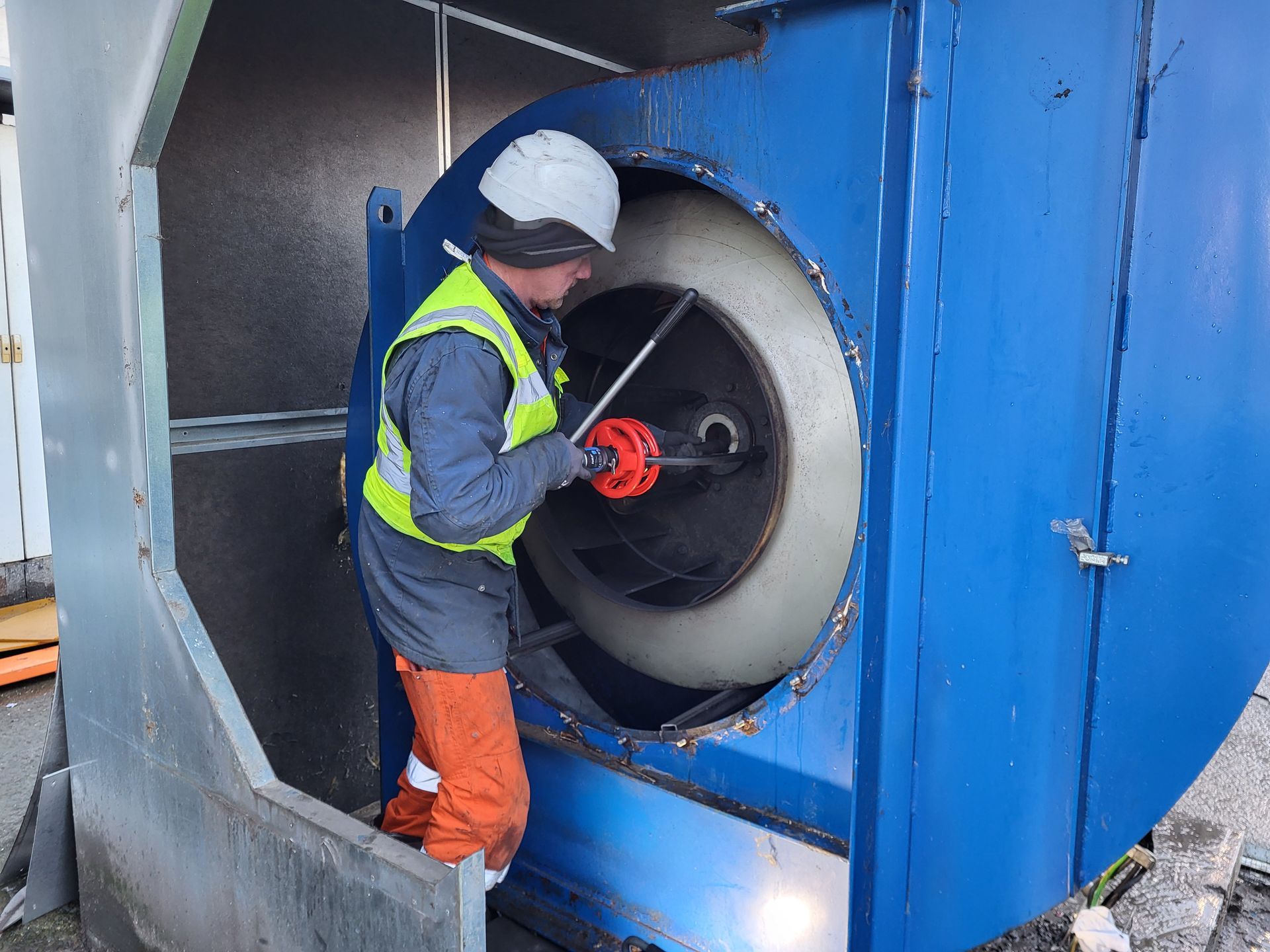 Een werknemer in veiligheidsuitrusting repareert een grote blauwe industriële ventilator in de buitenlucht.