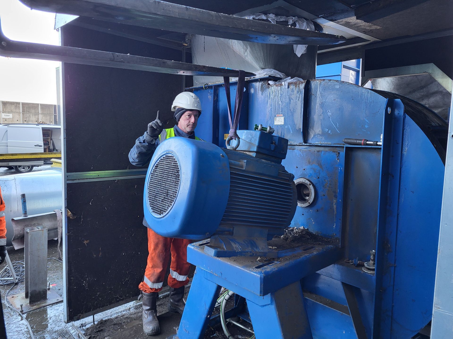 Een persoon in een oranje overall en veiligheidshelm is bezig een grote blauwe industriële motor af te stellen.