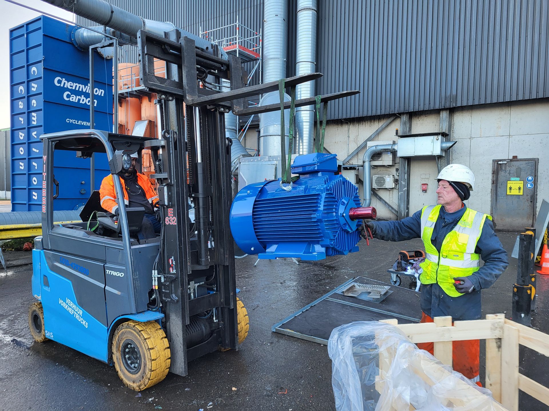 Werknemers gebruiken een heftruck om een grote blauwe industriële motor naar buiten te vervoeren.