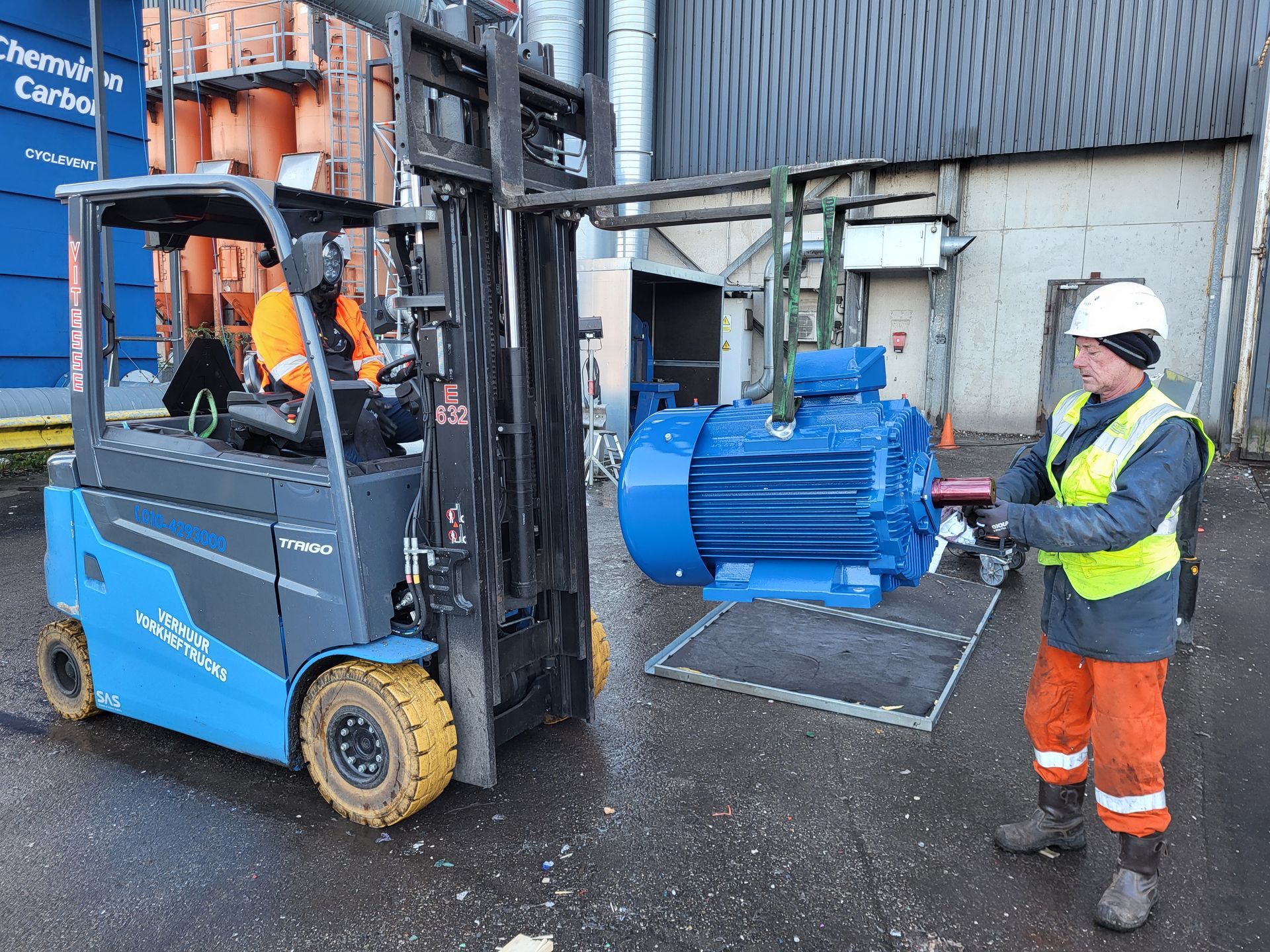 Een heftruck tilt een grote blauwe motor op; een medewerker in een veiligheidsvest begeleidt de motor