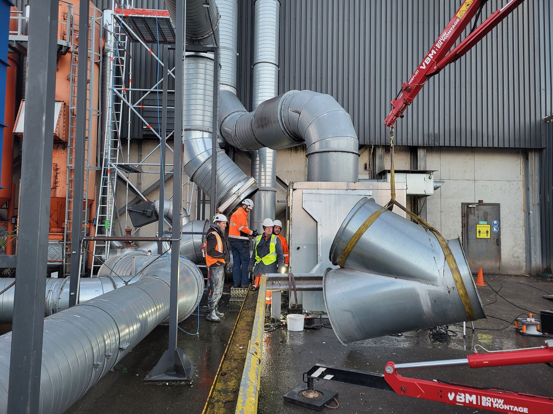 Arbeiders installeren grote metalen luchtkanalen in de buitenlucht, met behulp van een kraan.