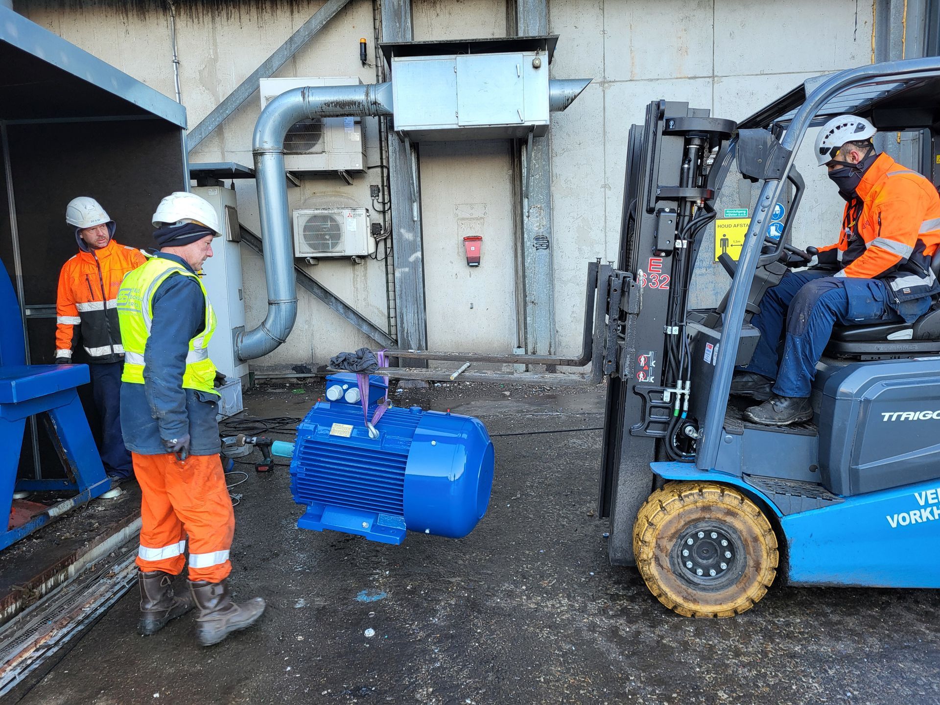 Werknemers in veiligheidsvesten gebruiken een heftruck om een grote blauwe motor in de buurt van een gebouw te verplaatsen.