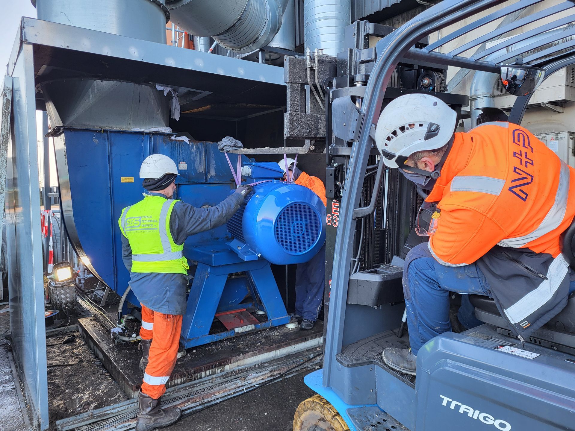 Arbeiders installeren een grote blauwe motor met een heftruck. Iedereen draagt veiligheidsuitrusting. Industriële omgeving.